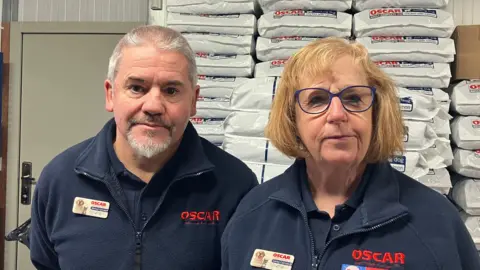 BBC Michael, left, has short grey hair with a grey goatee. He is wearing a navy fleece with red writing saying Oscar. Lesley, right, has short ginger hair and is also wearing the same navy fleece.