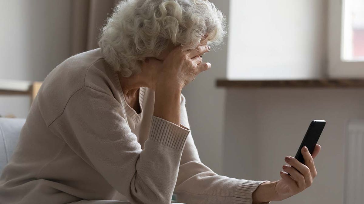 Sad elderly woman sit on sofa hold smartphone feels disappointed by received sms bad news, awful message, difficulties with modern device usage, unpleasant notification, stressed older person concept