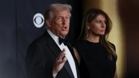 Getty Images President Donald Trump and first lady Melania Trump pose on the red carpet for the 2025 Kennedy Center Honors. Trump, in a tuxedo, tooks towards the camera as he gesture while speaking to reporters 